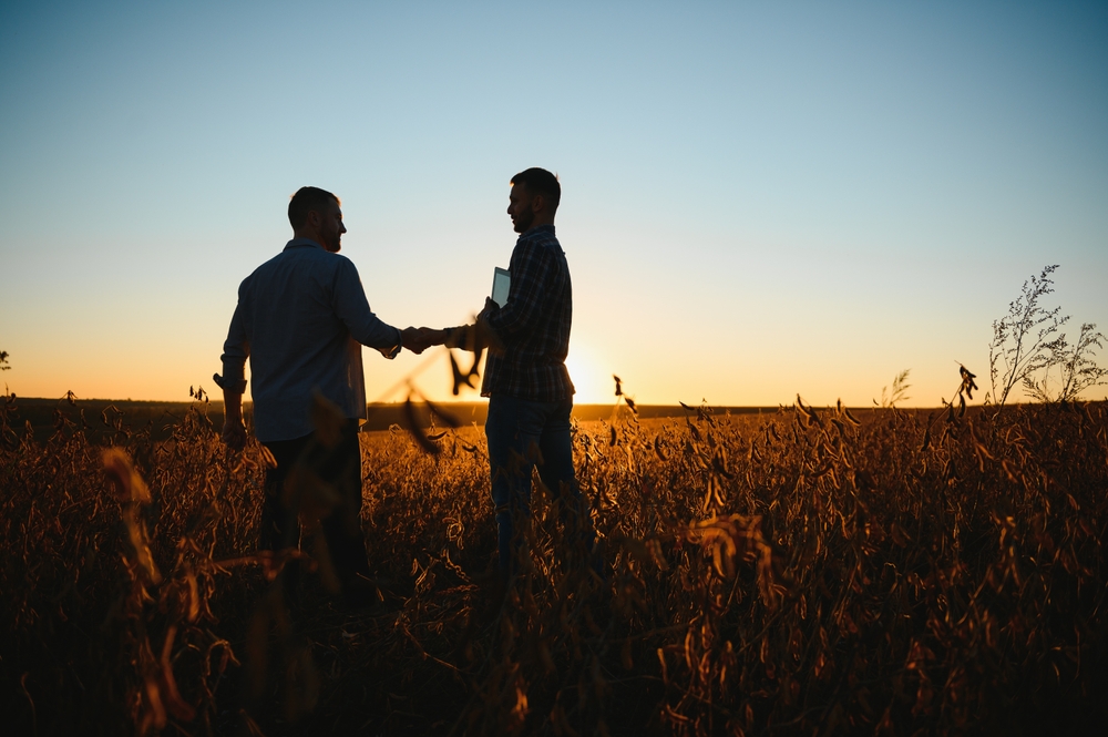 Two people shaking hands at sunset in a soybean field, one holding a tablet